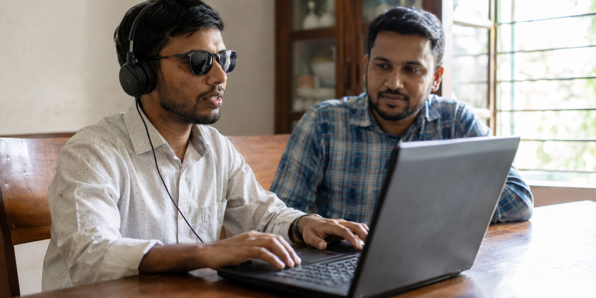 Bangladeshi man with visual impairment using a laptop with headphones while receiving guidance